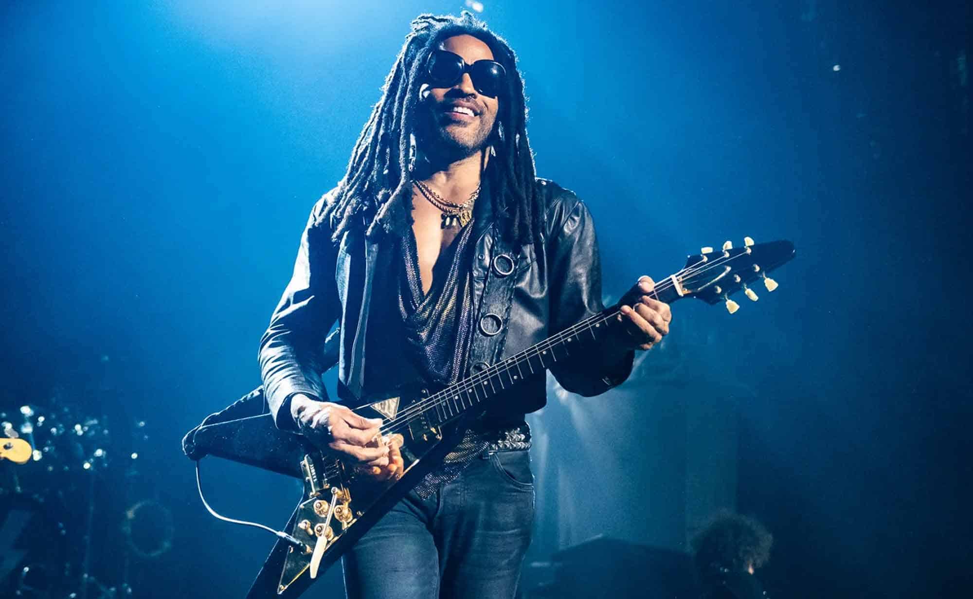 An energetic musician with dreadlocks, sunglasses, and leather jacket performing on stage with electric guitar and dynamic stage lighting.