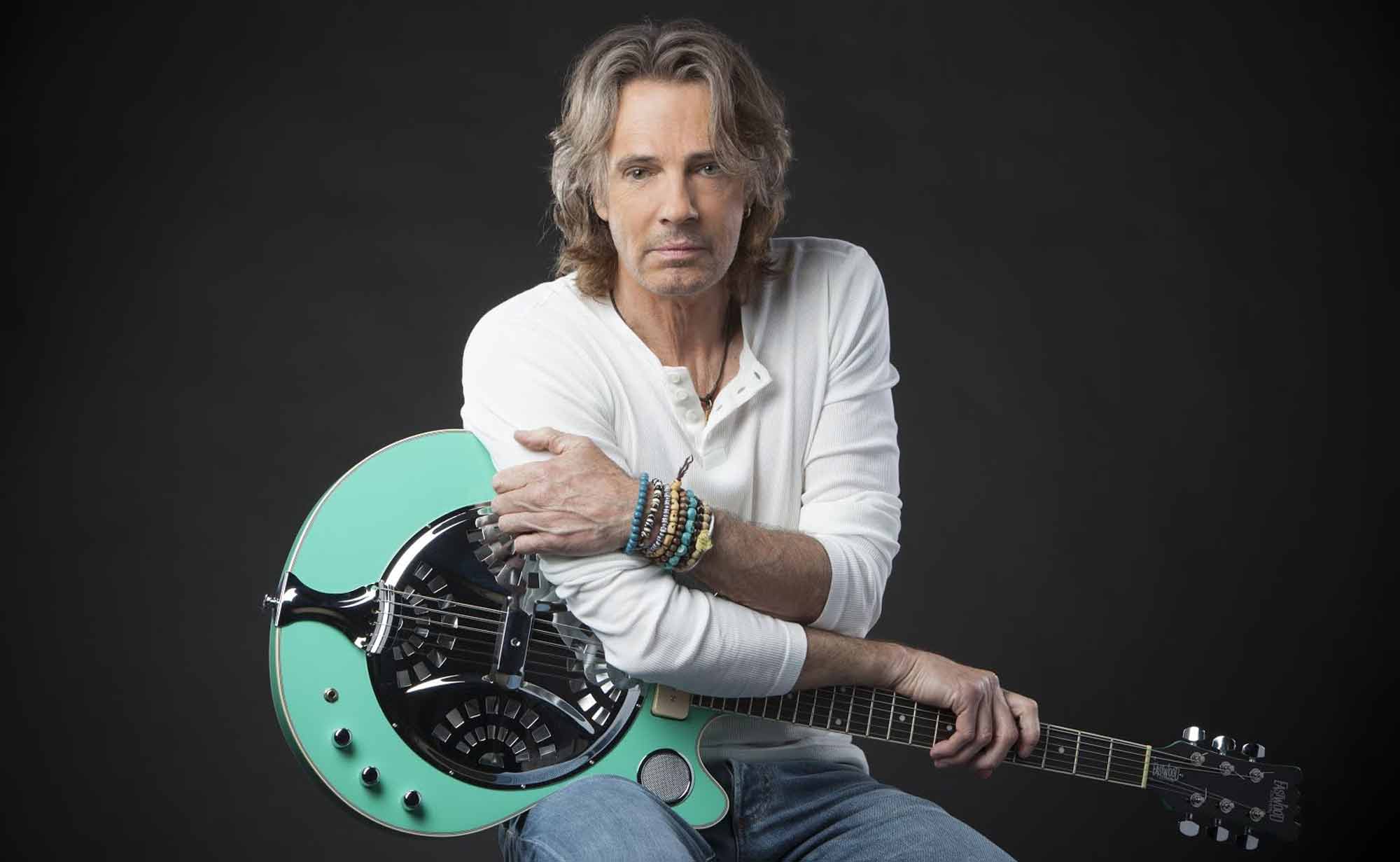 A mature male musician with long hair and bracelets, holding a stylish electric guitar against a dark backdrop.