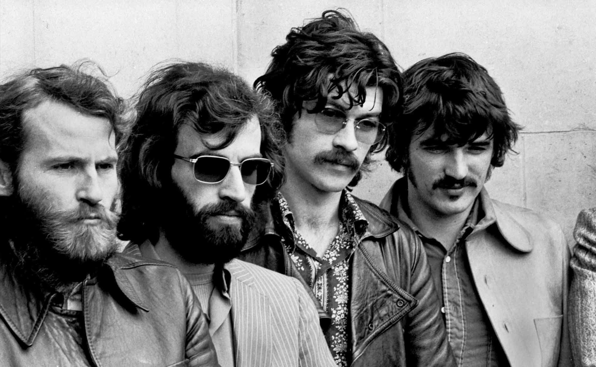 Older black and white photo of a rock band with four men in leather jackets, sunglasses, and stylish hairstyles, from the late 20th-century music scene.