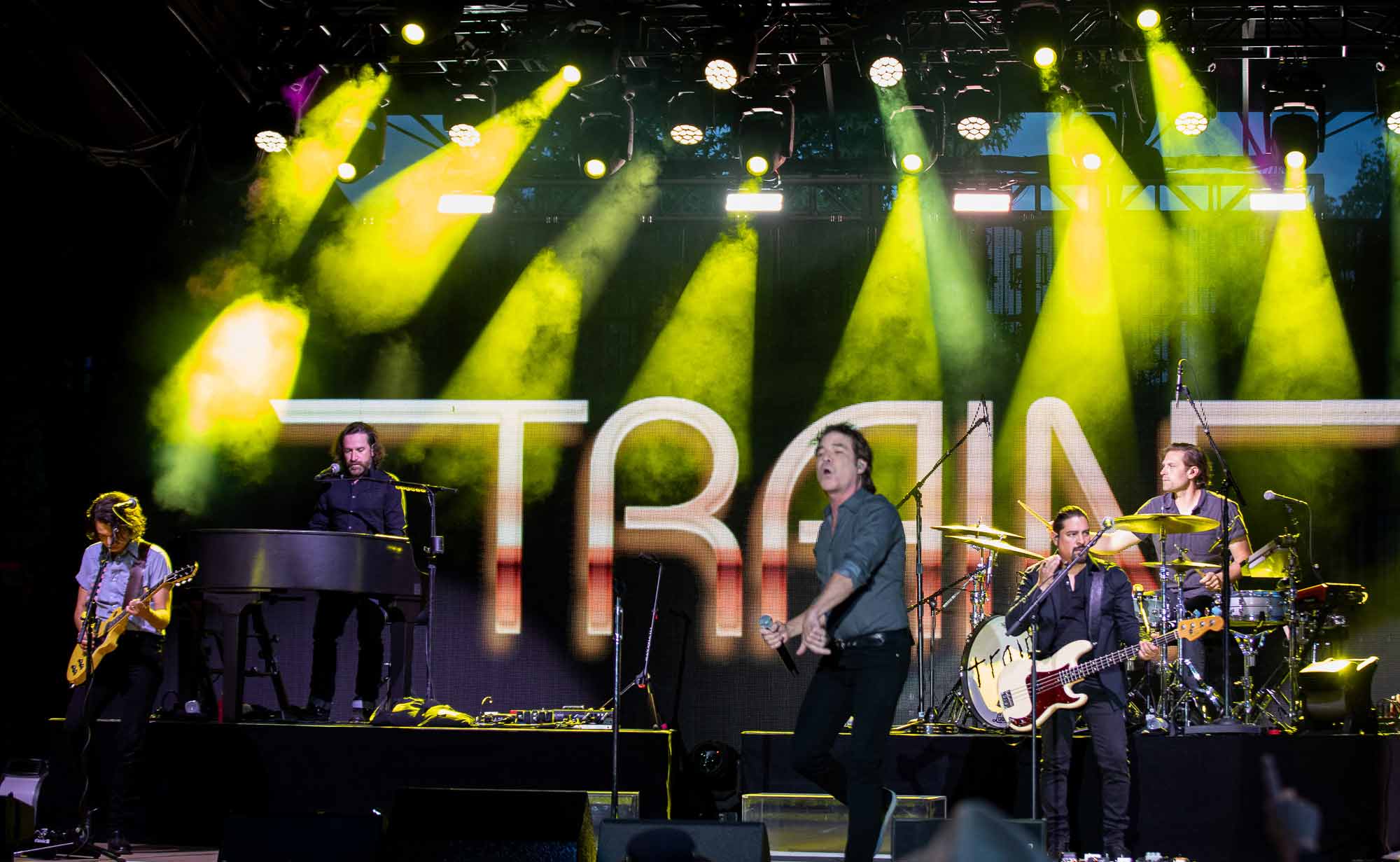 Energetic live band performing on outdoor stage with bright yellow lights and dynamic concert atmosphere.