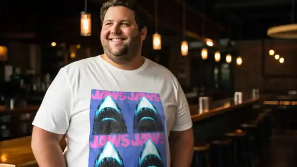 A smiling man stands in a warmly lit bar, wearing a Jaws Jaws graphic T-shirt, creating a casual and inviting atmosphere.