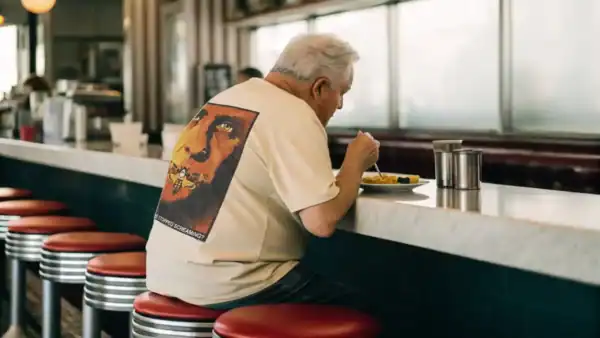 An elderly man with white hair eats alone at a retro diner counter, sitting on red stools, enjoying a meal on a sunny day.