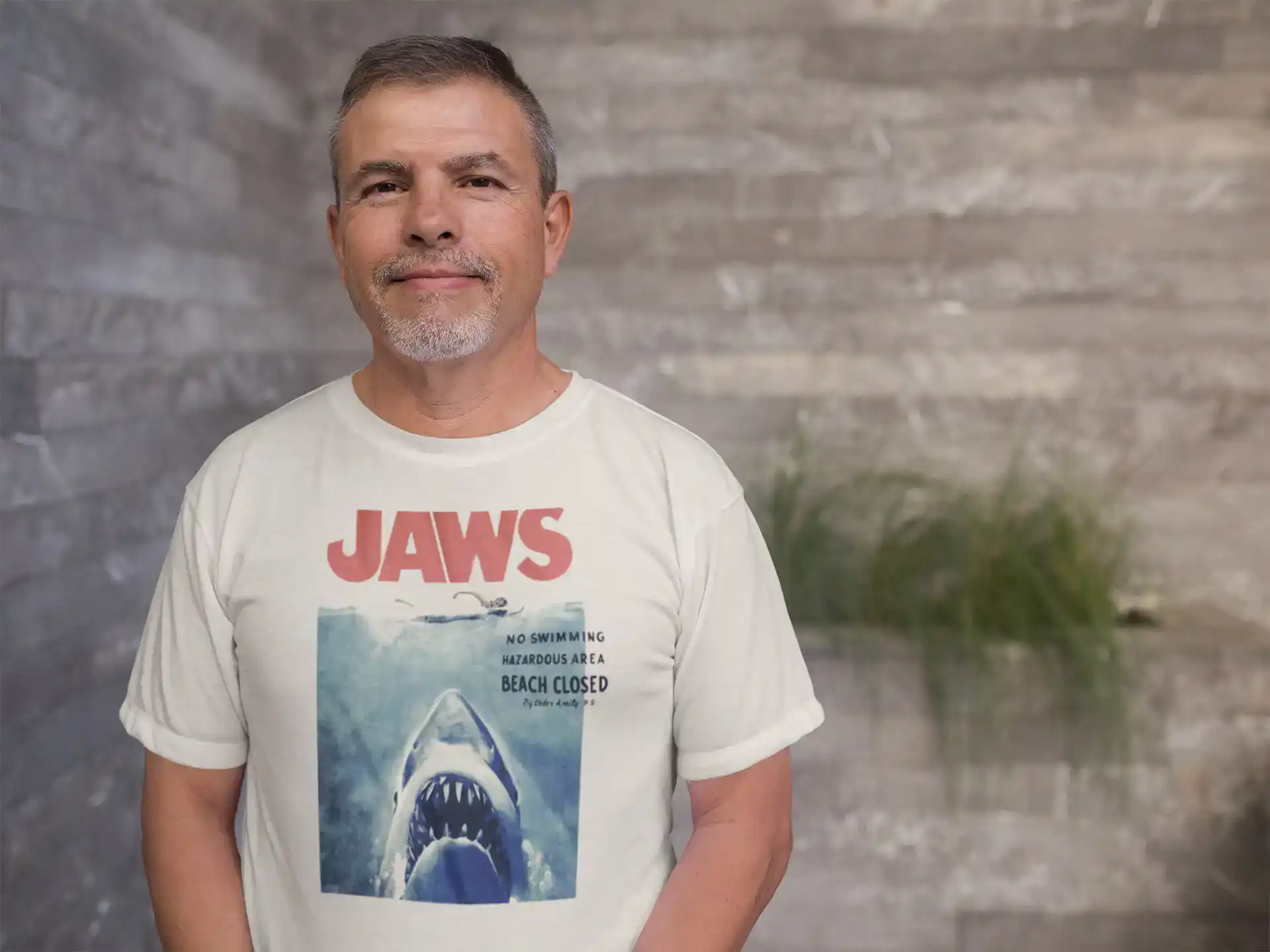 A middle-aged man stands confidently in a casual Jaws t-shirt against a modern rustic background.