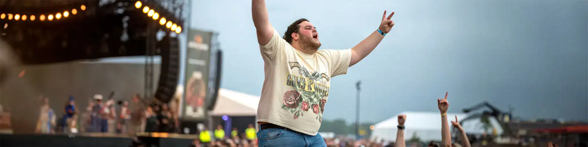 A lively crowd at an outdoor concert, with a man in a graphic t-shirt dancing and raising his hand, surrounded by festival lights and stages.