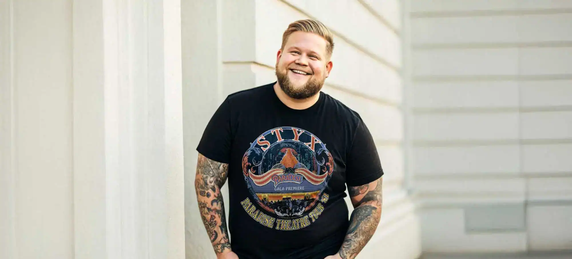 A cheerful man with tattoos smiling, wearing a black band T-shirt featuring Styx and Paradise Theatre Tour, standing against a modern building wall.