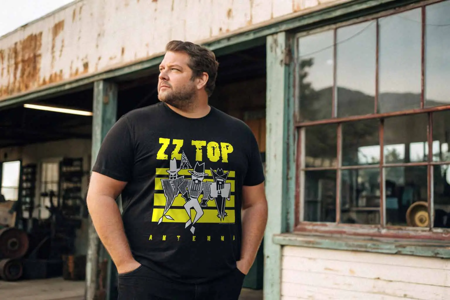 A man wearing a ZZ Top concert tee standing outdoors in front of an old, weathered building with rustic charm and large windows, emphasizing retro and rock style.