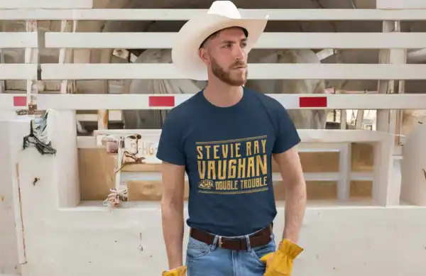A man in a cowboy hat and graphic tee with "Stevie Ray Vaughan Double Trouble" stands against a rustic fence with cows in the background.