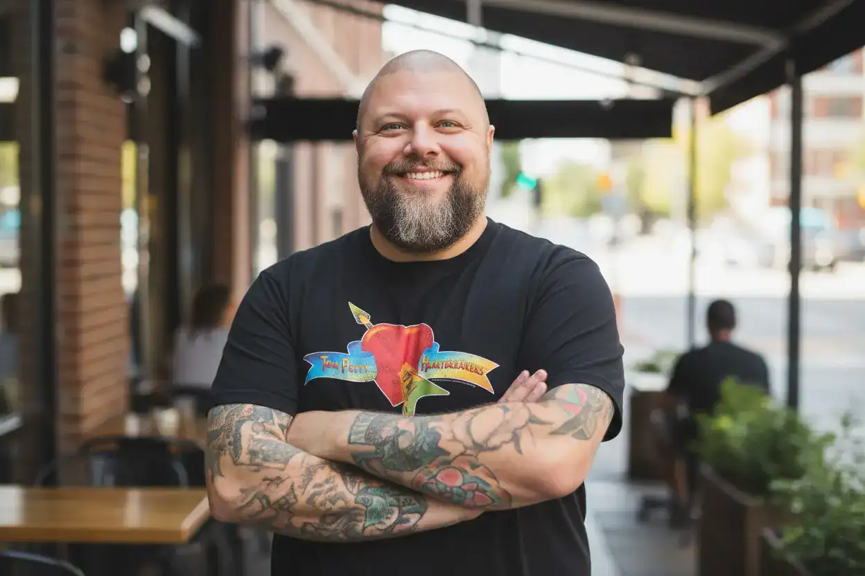 A smiling man with tattoos stands confidently outdoors at a cafe, showcasing his casual style on a sunny day.
