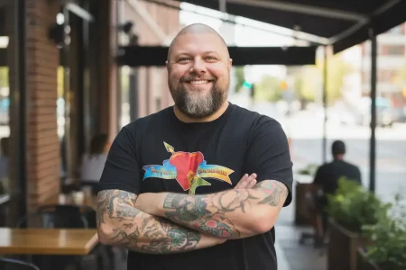 A smiling man with tattoos stands confidently outdoors at a cafe, showcasing his casual style on a sunny day.