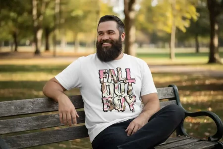 A man with a beard smiles while sitting on a park bench during fall, wearing a "Fall Out Boy" graphic T-shirt, surrounded by colorful autumn trees and a peaceful outdoor setting.