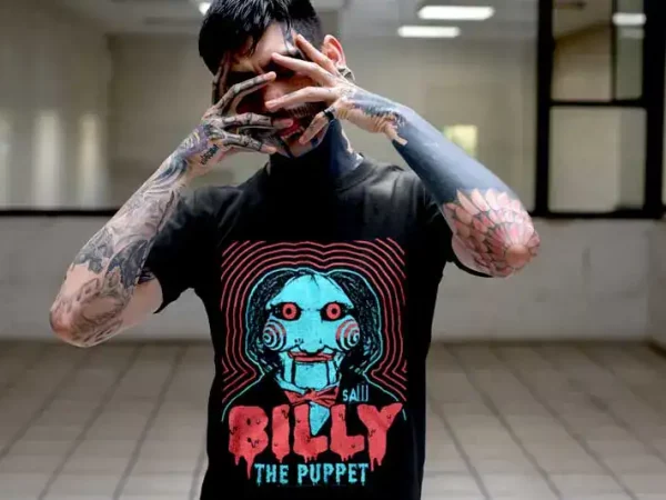 A person wearing a Billy Puppet horror-themed graphic T-shirt, showcasing unique tattoo art on arms, in an urban setting.