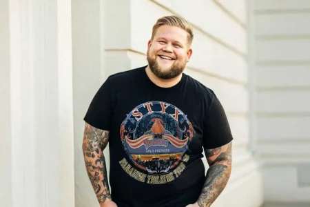 A cheerful man with tattoos smiling, wearing a black band T-shirt featuring Styx and Paradise Theatre Tour, standing against a modern building wall.